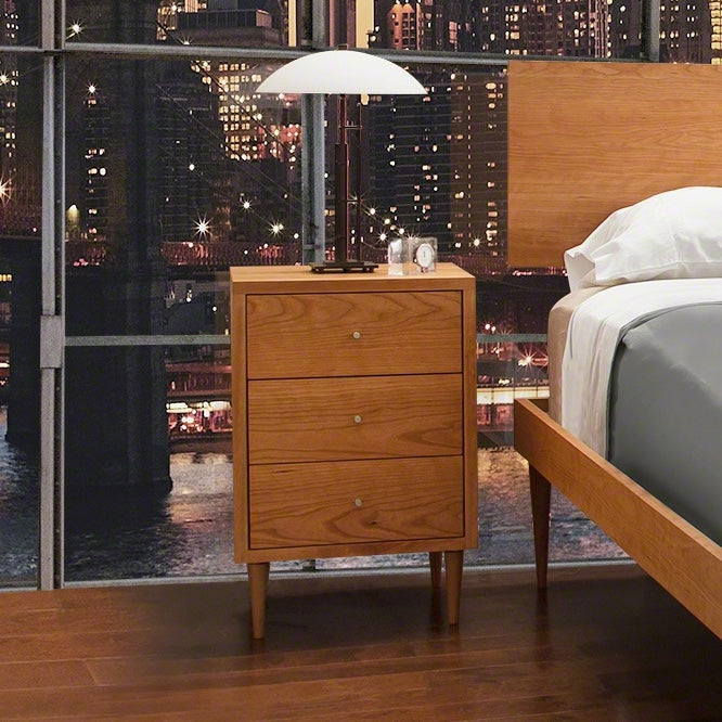 A wooden nightstand with three drawers beside a bed, overlooking a cityscape through a floor-to-ceiling window at night. a lamp and a small clock sit atop the nightstand.