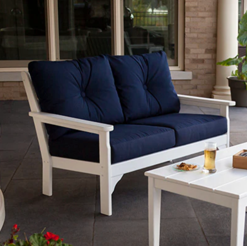 White wooden outdoor loveseat with deep blue cushions on a patio, accompanied by a small white side table with a snack and a drink on it.