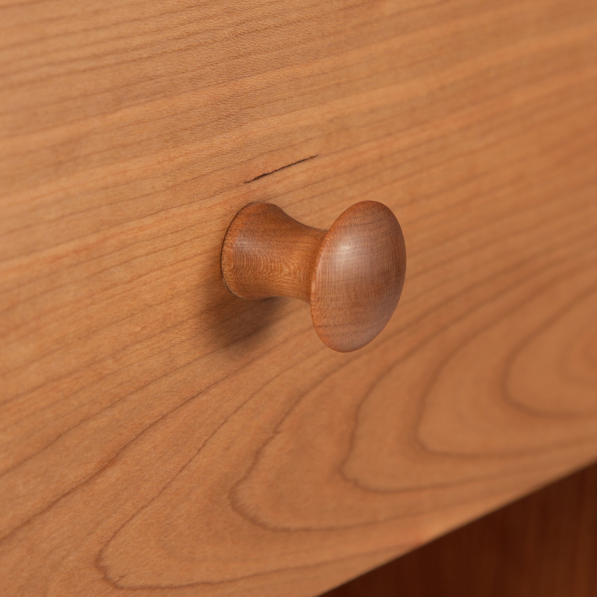 Close-up of a Maple Corner Woodworks American Shaker 1-Drawer Enclosed Shelf Nightstand knob on the smooth, grainy surface, showcasing a natural cherry wood finish and Vermont craftsmanship.