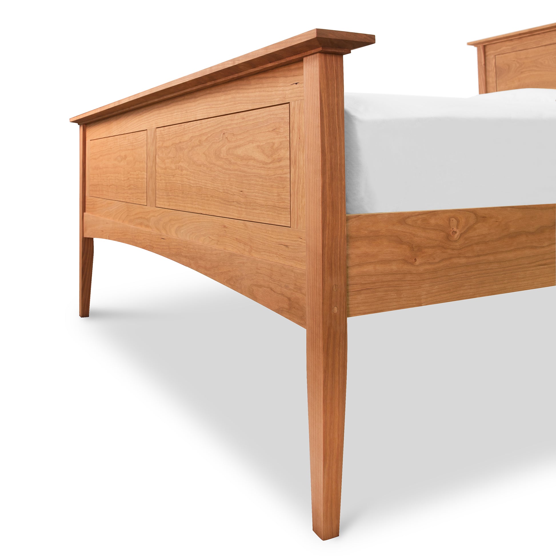 A wooden bed frame, handmade from sustainably harvested woods, with a prominent headboard featuring Shaker panel details and simple, streamlined legs, set against a white background. The bed is partially covered by the American Shaker Panel Bed by Maple Corner Woodworks.