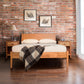 A contemporary bedroom with a mid-century modern Maple Corner Woodworks Andover Modern Incline Bed and two nightstands against an exposed brick wall. A plaid throw drapes over the bed. Two framed sketches hang above the bed, and