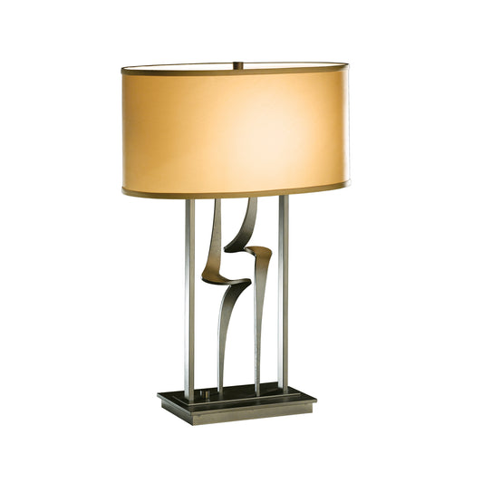 The Hubbardton Forge Antasia #1 Table Lamp features a hand-forged iron base with unique wavy patterns and a cylindrical, golden shade that emits a warm glow. It stands on a square, flat base.