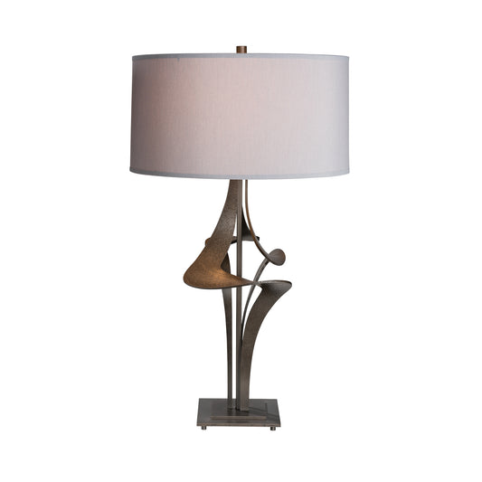 An Hubbardton Forge Antasia #2 Table Lamp featuring a stylized metal base with smooth curves and two abstract shapes, topped with a large, cylindrical light grey shade.
