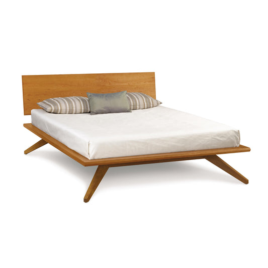 A Copeland Furniture mid-century modern-contemporary style Astrid Cherry Platform Bed frame with a solid cherry wood headboard and angled legs, dressed with a white fitted sheet and adorned with two striped and one solid-colored throw pillows.