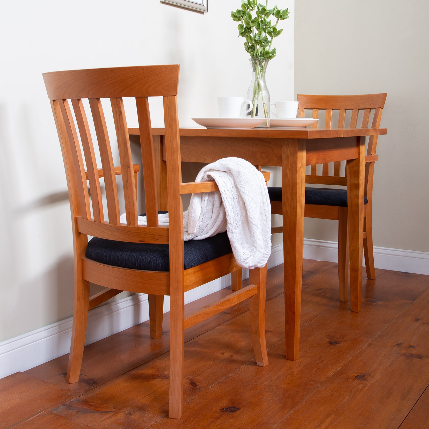 Cherry wood table paired with Lyndon Furniture's Bistro Dining Chairs, featuring one chair adorned with a white cloth and a vase centered on the tabletop.