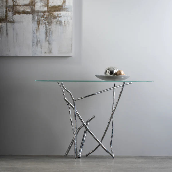 A modern console table with a glass top and an abstract, metallic branch-like base is placed against a plain wall. A metal bowl containing metallic spheres is centered on the table. An abstract painting with neutral tones is partially visible on the wall above.