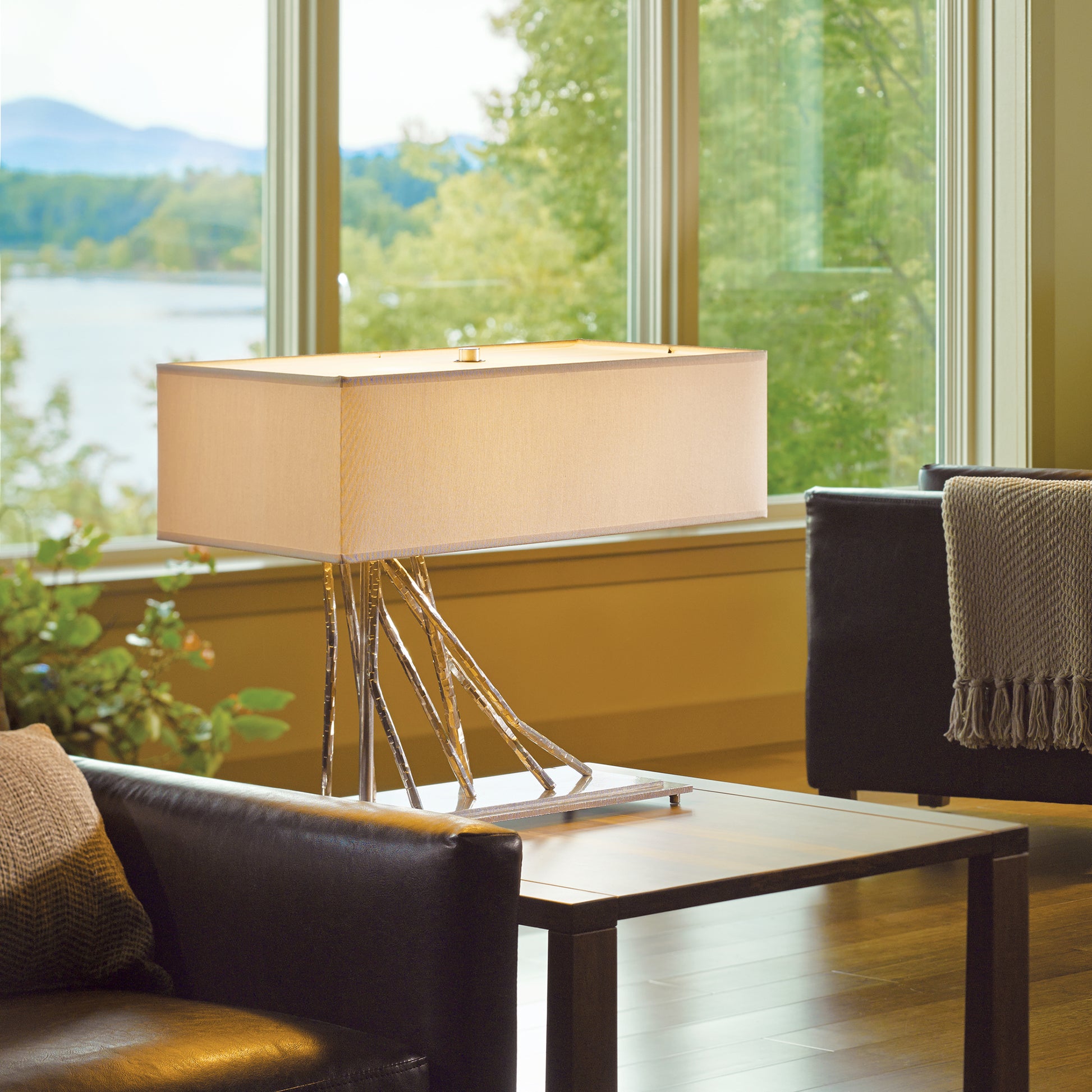 A contemporary room with a large window overlooking a scenic lake. A Hubbardton Forge Brindille Table Lamp with a unique metal base and rectangular shade stands on a small table between two dark leather chairs. A woven throw drapes