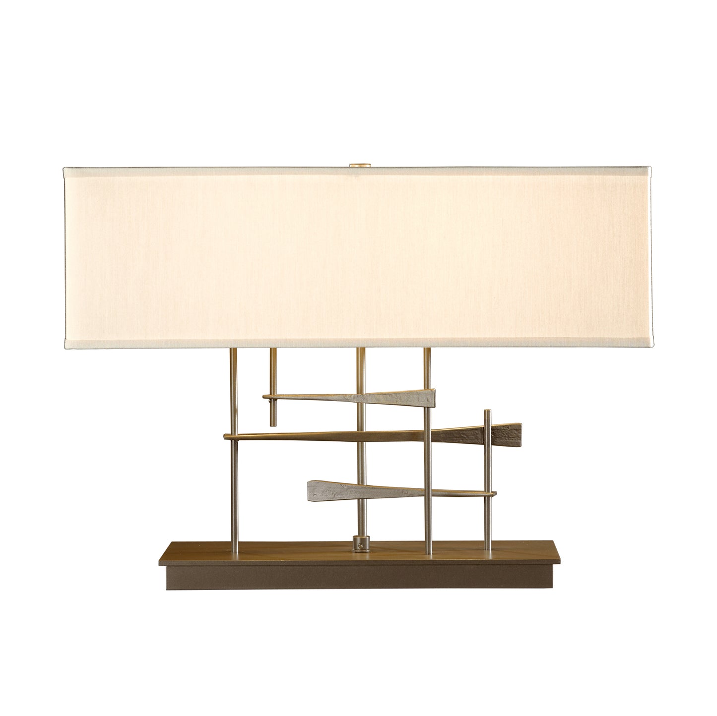 A modern Cavaletti table lamp with a rectangular off-white shade and a hand-forged wrought iron base in a gold and brown geometric pattern, isolated on a white background by Hubbardton Forge.