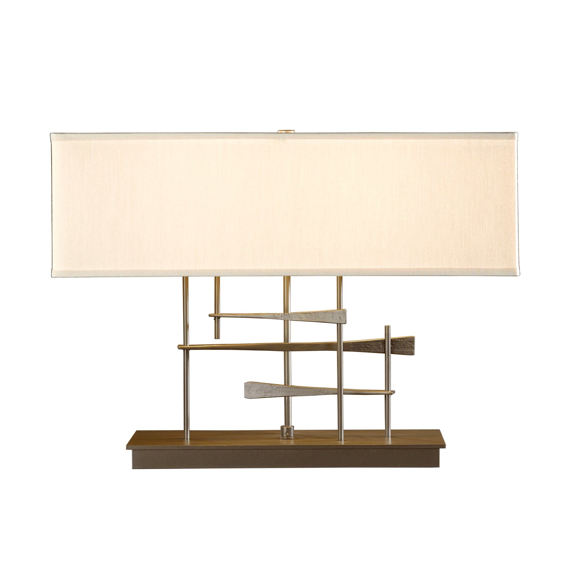 A modern Cavaletti table lamp with a rectangular off-white shade and a hand-forged wrought iron base in a gold and brown geometric pattern, isolated on a white background by Hubbardton Forge.