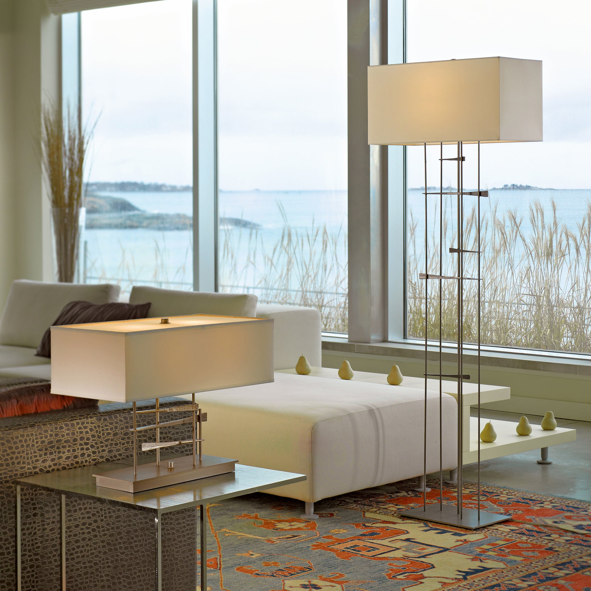 Modern living room overlooking a beach, featuring a white sofa, Hubbardton Forge Cavaletti Table Lamp, side table with books, and a colorful area rug, all bathed in natural light from large windows.