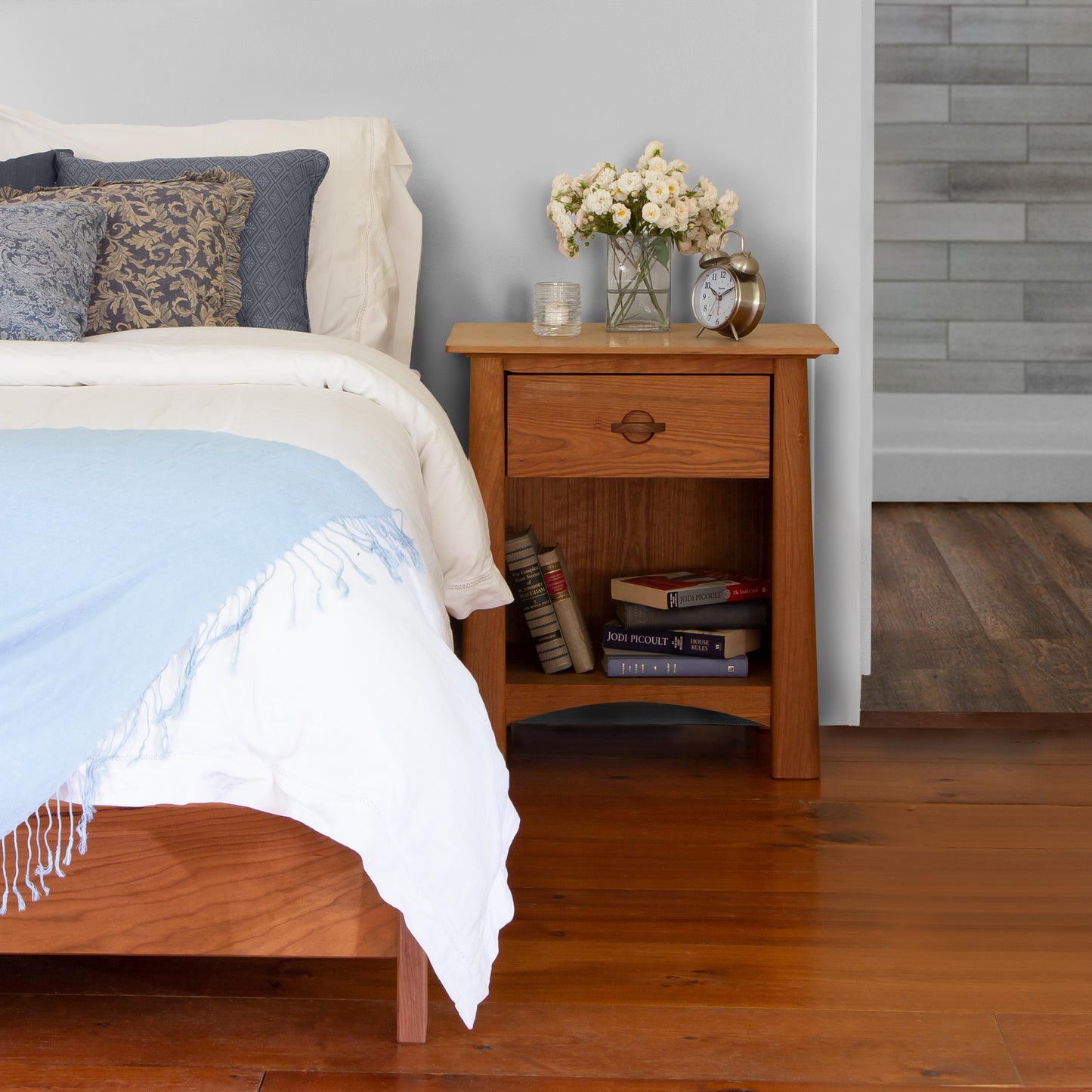 A neatly made bed with a blue and white blanket, beside a Maple Corner Woodworks Cherry Moon 1-Drawer Enclosed Shelf Nightstand holding flowers, books, and a clock, in a room with sustainably harvested hardwood flooring.