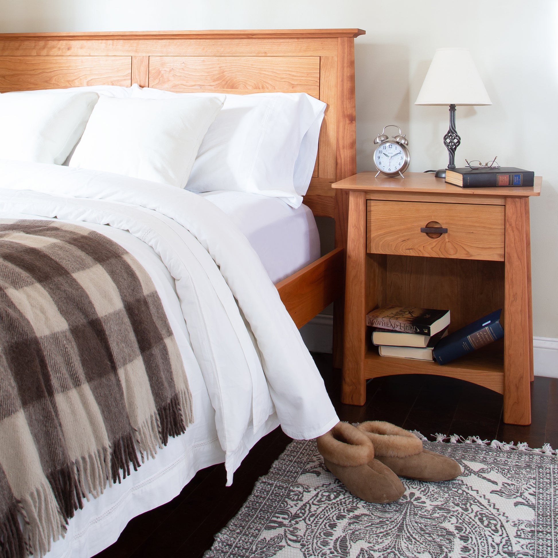 Wooden bed with white sheets and plaid blanket, accompanied by a Cherry Moon 1-Drawer Enclosed Shelf Nightstand by Maple Corner Woodworks, crafted from sustainably harvested hardwoods.