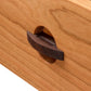 Close-up of a wooden drawer from the Cherry Moon 7-Drawer Dresser by Maple Corner Woodworks with a dark brown, uniquely shaped handle. The wood grain is visible and provides a natural texture on the surface.