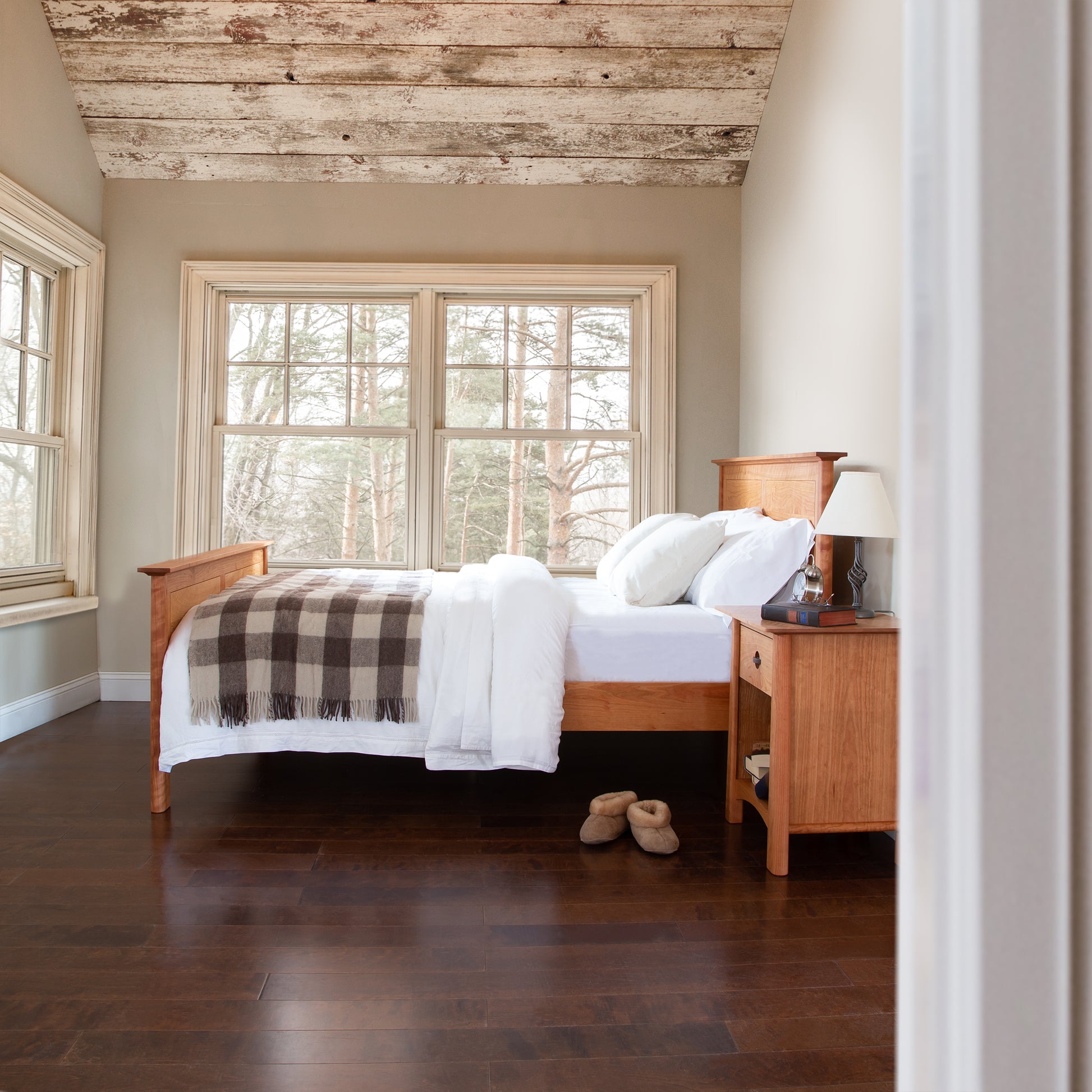 A cozy bedroom with a Maple Corner Woodworks Cherry Moon Panel Bed and side table, white bedding, and a plaid throw. Slippers are placed on the hardwood floor, large windows show a wooded view, and the