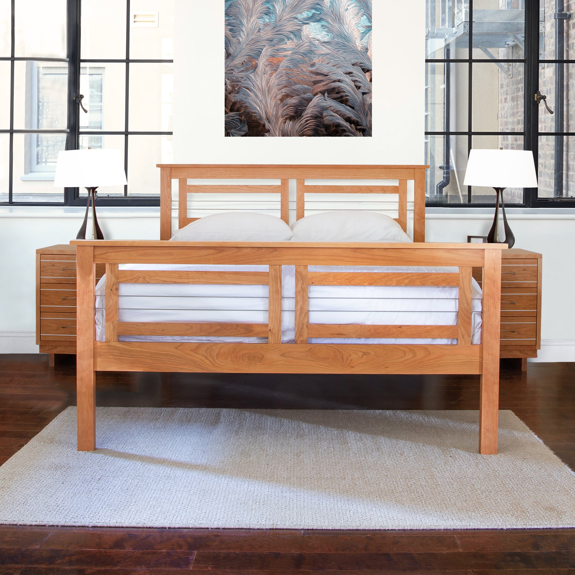 A solid hardwood construction bed frame with white bedding is situated on a cream rug in a room with large windows and a framed abstract wall art above the Vermont Furniture Designs Contemporary Cable Bed.