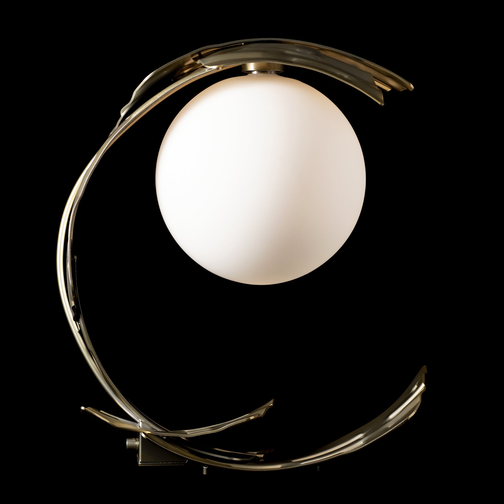 Spherical white bulb encased in an ornate, curved brass frame of the Hubbardton Forge Crest Table Lamp.
