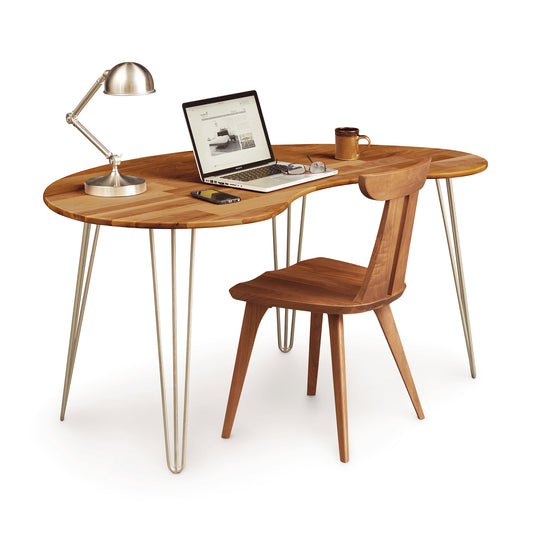 A solid natural walnut Copeland Furniture Essentials Kidney Shaped Desk with metal legs, equipped with a laptop, a coffee cup, and a desk lamp, accompanied by a matching wooden chair on a white background.