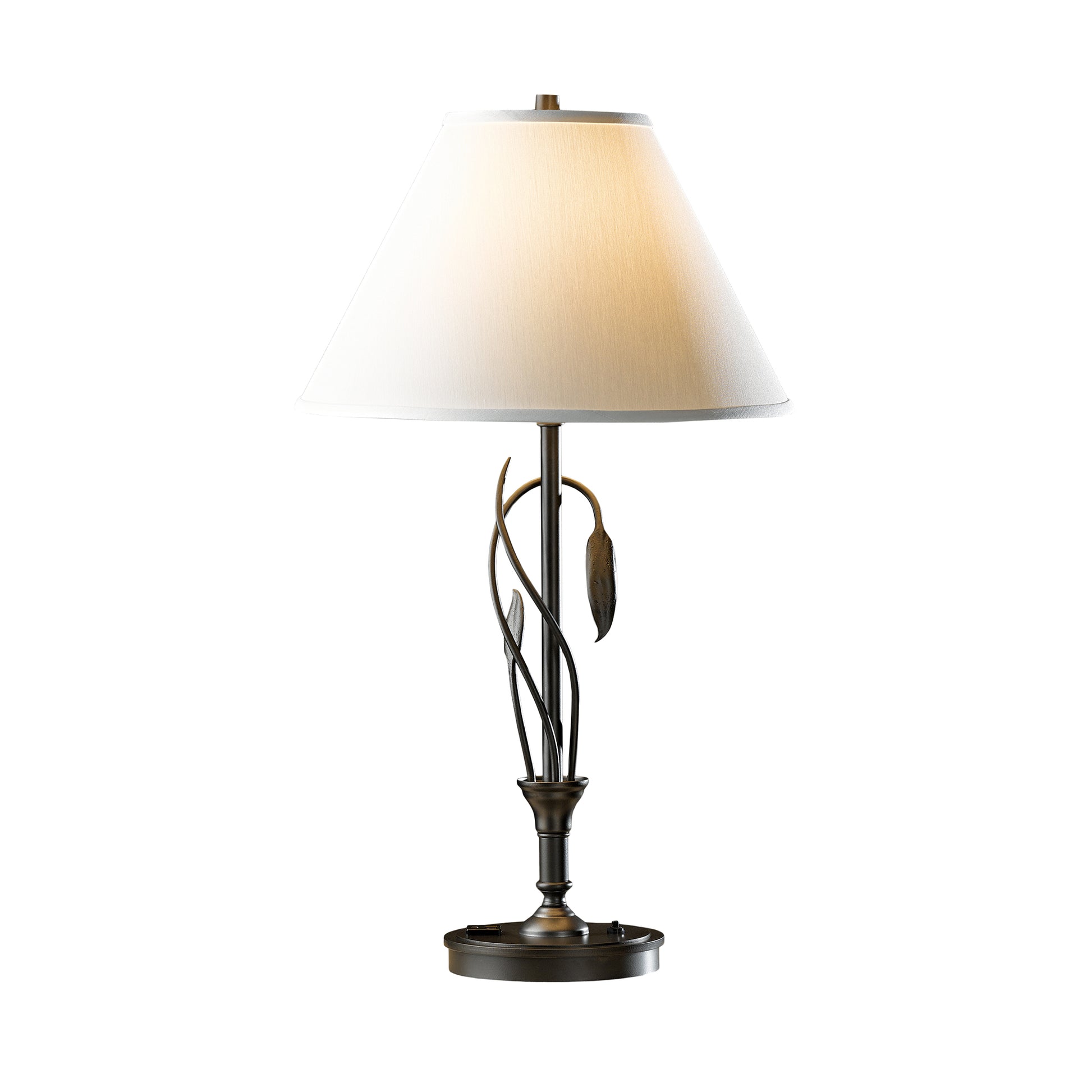 A Forged Leaves and Vase Table Lamp by Hubbardton Forge with a hand-crafted bronze base featuring leaf motifs and twisted stems, supporting a white conical lampshade, illuminated against a white background.
