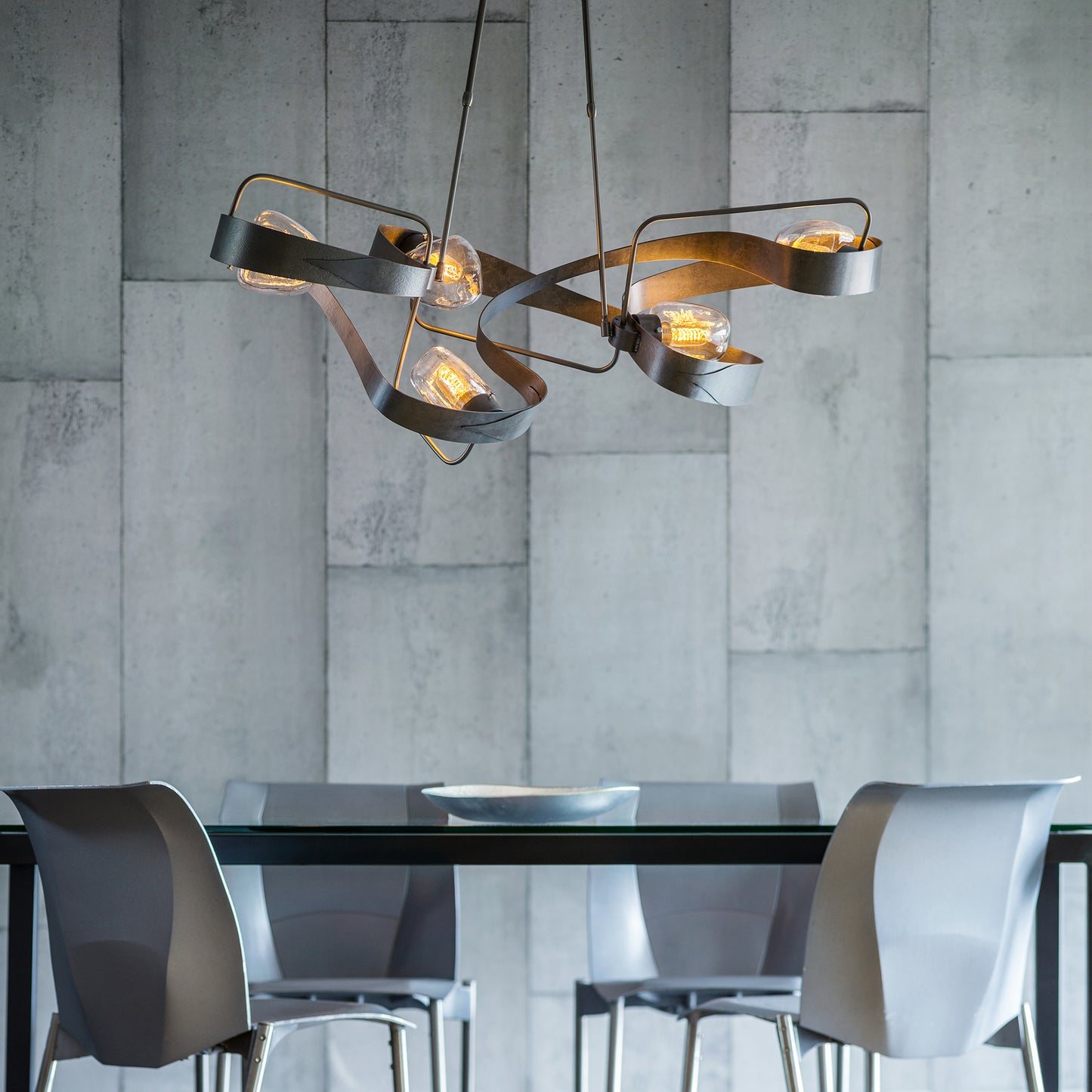 Graffiti Pendant by Hubbardton Forge, a striking modern metal light fixture, suspended above a sleek glass table paired with four elegant gray chairs.