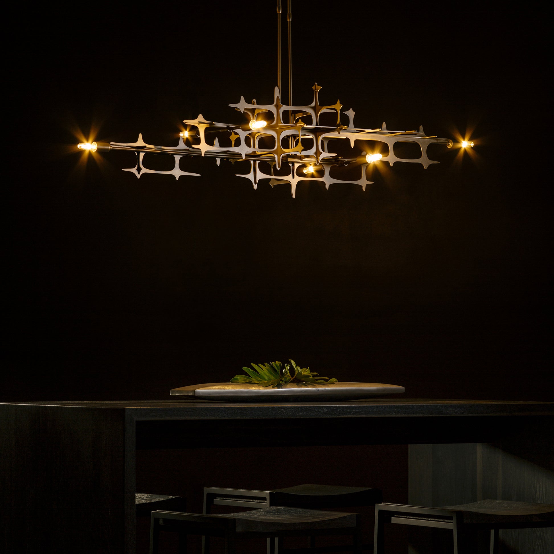 An industrial design Grid Pendant chandelier by Hubbardton Forge gracefully hangs over a table in a dark room.