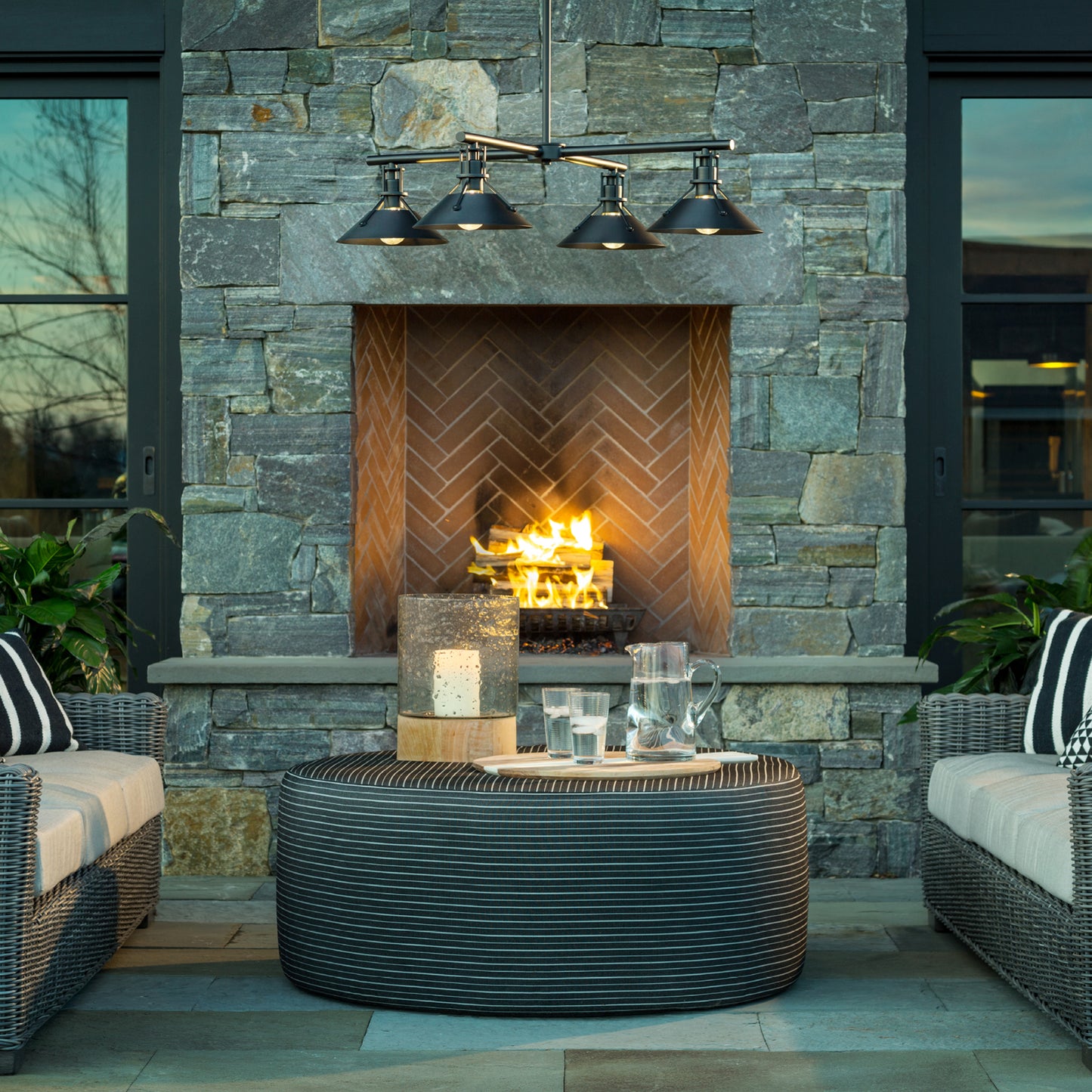 Round table with striped-cushion seating and Henry Outdoor 4-Light Pendant by Hubbardton Forge, highlighting elegant outdoor furniture.