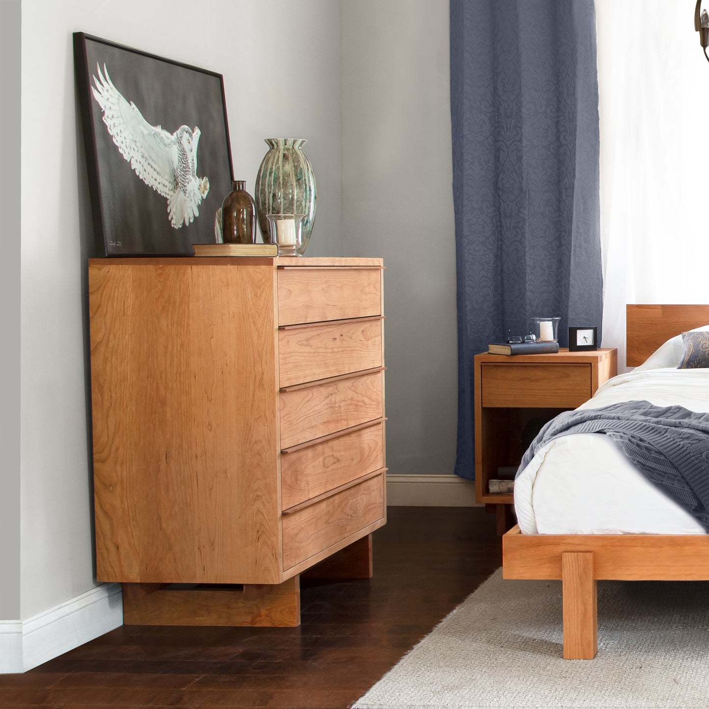 A handcrafted wooden dresser, known as the Vermont Furniture Designs Kipling 5-Drawer Wide Chest, stands in a bedroom with a framed artwork above it, next to a window draped with blue curtains.