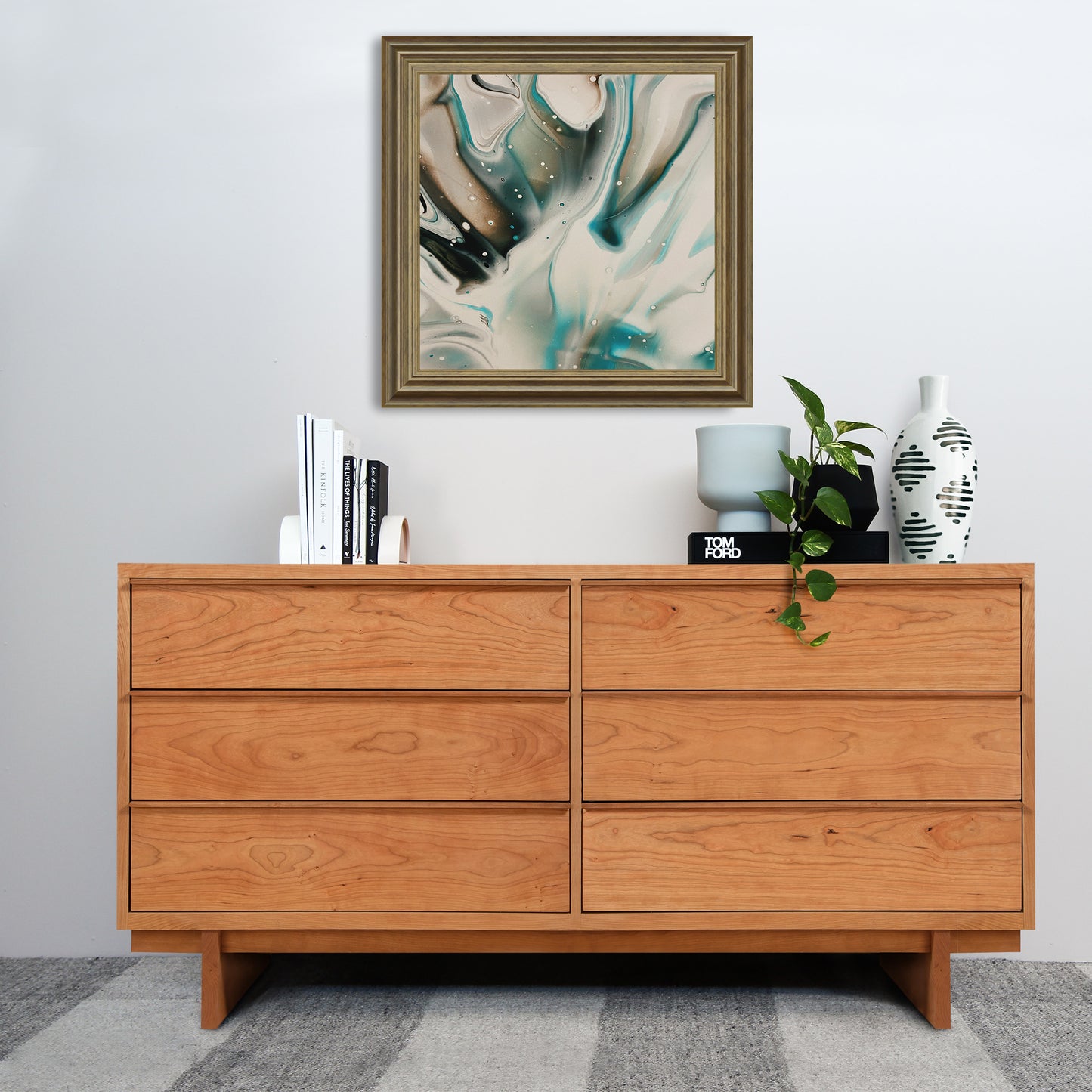 A modern design Vermont Furniture Designs Kipling 6-Drawer Dresser in natural cherry stands against a white wall, topped with decorative items including a framed abstract painting, books, a clock, and plant pots.