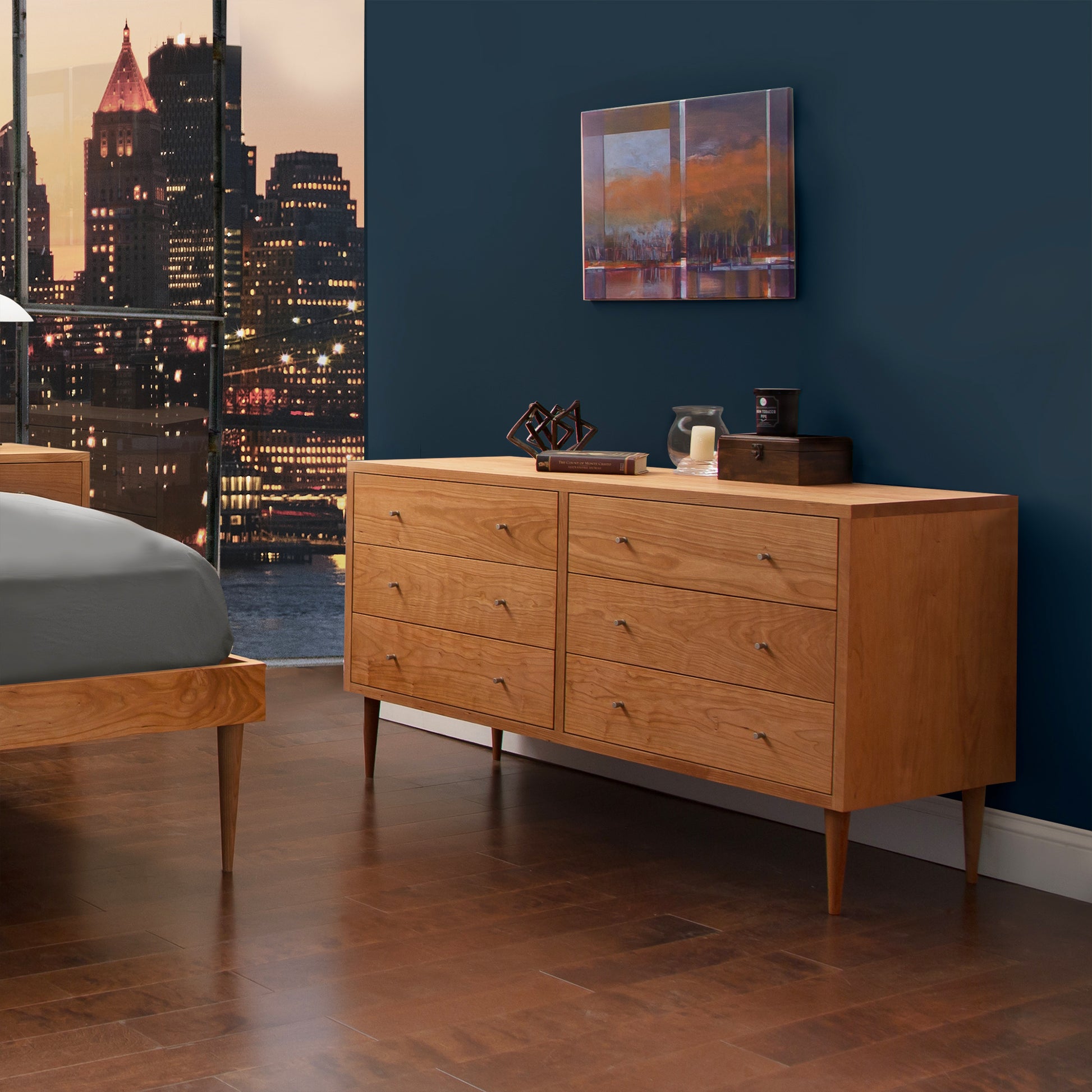 A modern bedroom featuring a Vermont Furniture Designs Larssen 6-Drawer Dresser of solid wood construction and mid-century modern design with six drawers, next to which is a smaller bedside table with a lamp and a cup, against a