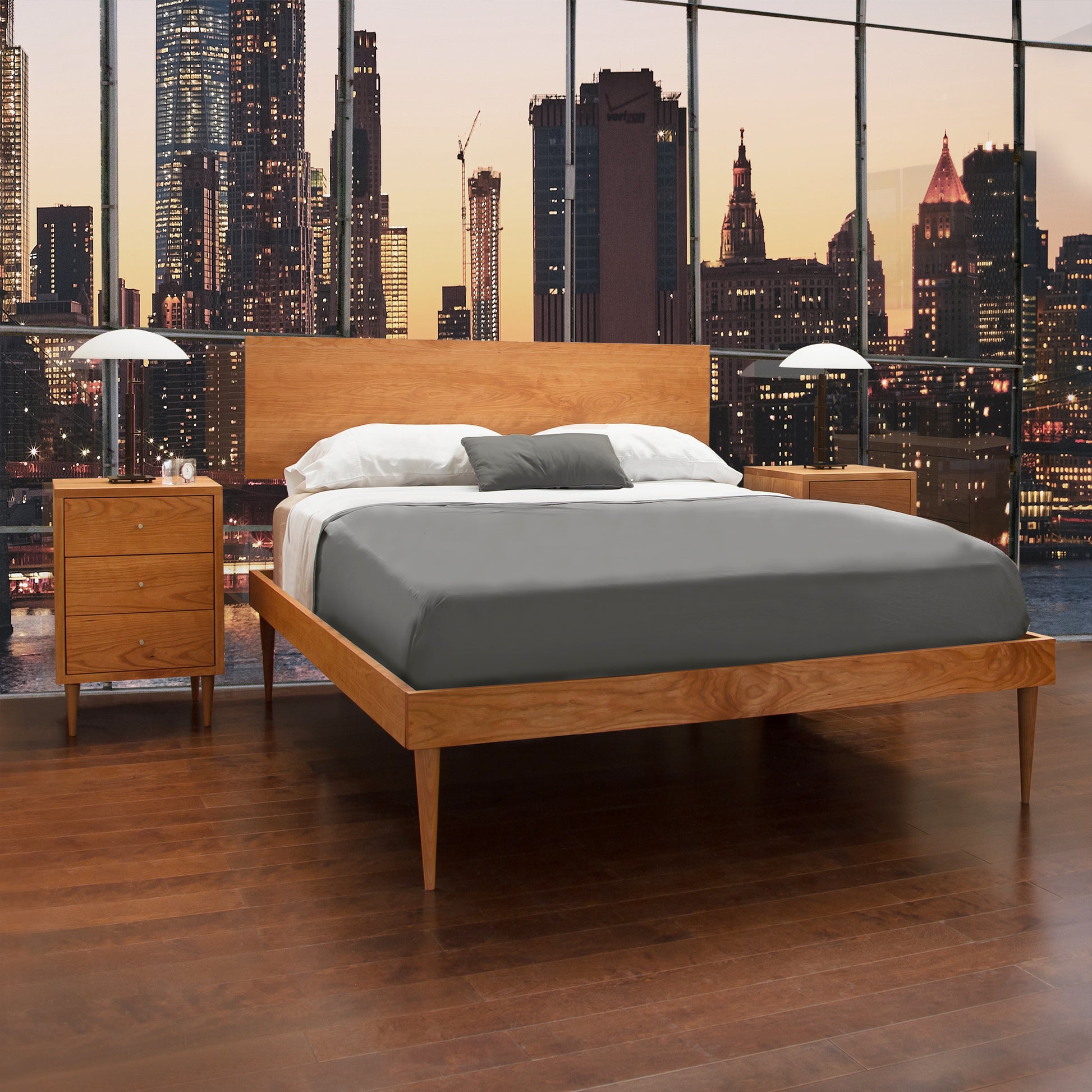 Mid-century modern design natural cherry Vermont Furniture Designs Larssen Bed with white bedding against a large window overlooking a city skyline at dusk.