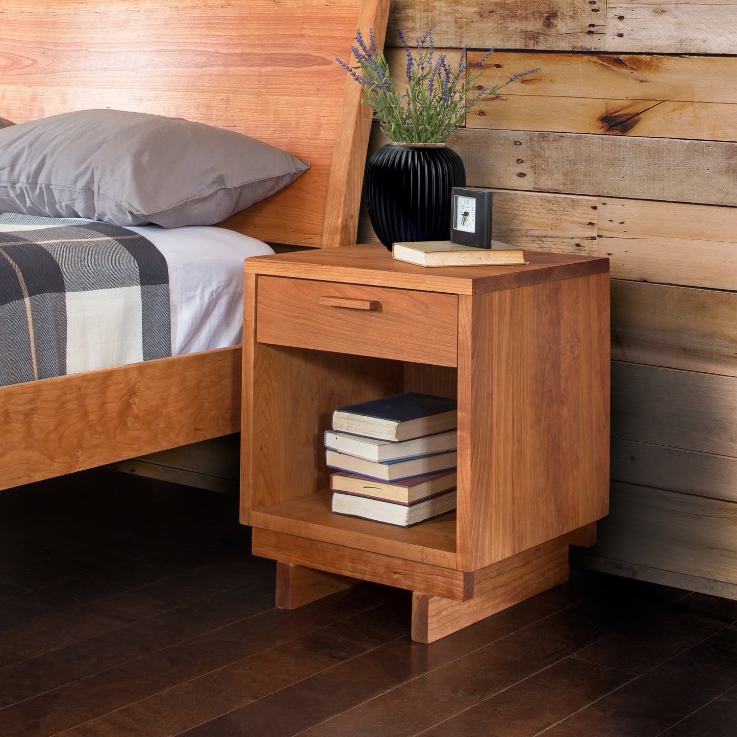 A Vermont Furniture Designs Loft 1-Drawer Enclosed Shelf Nightstand with an open drawer and a lower shelf holding a stack of books, located next to a bed with plaid bedding. A black vase with lavender flowers and a small