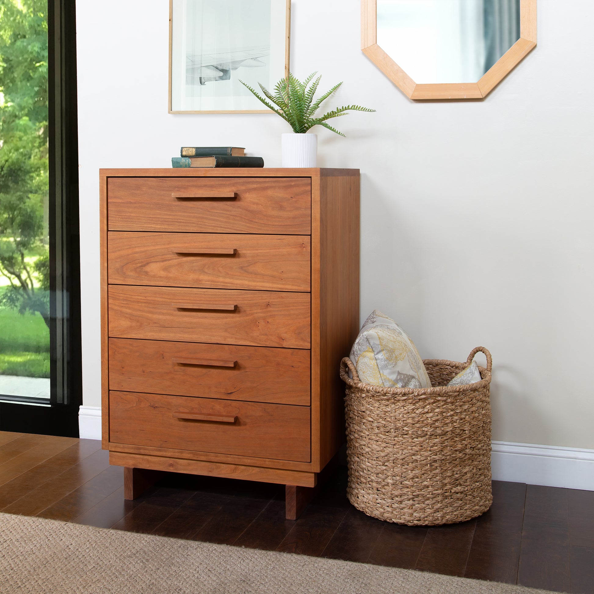 A solid hardwood Vermont Furniture Designs Loft 5-Drawer Chest stands next to a large window, with a woven basket containing pillows placed alongside it. On top of the chest, there is a small plant and a stack