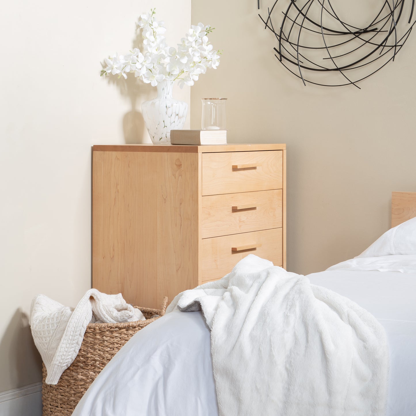 A modern bedroom corner featuring a Vermont Furniture Design Loft 5-Drawer Chest, exemplifying fine Vermont craftsmanship and made from solid hardwood, a white vase with white flowers on top, and an abstract black wall decor above.