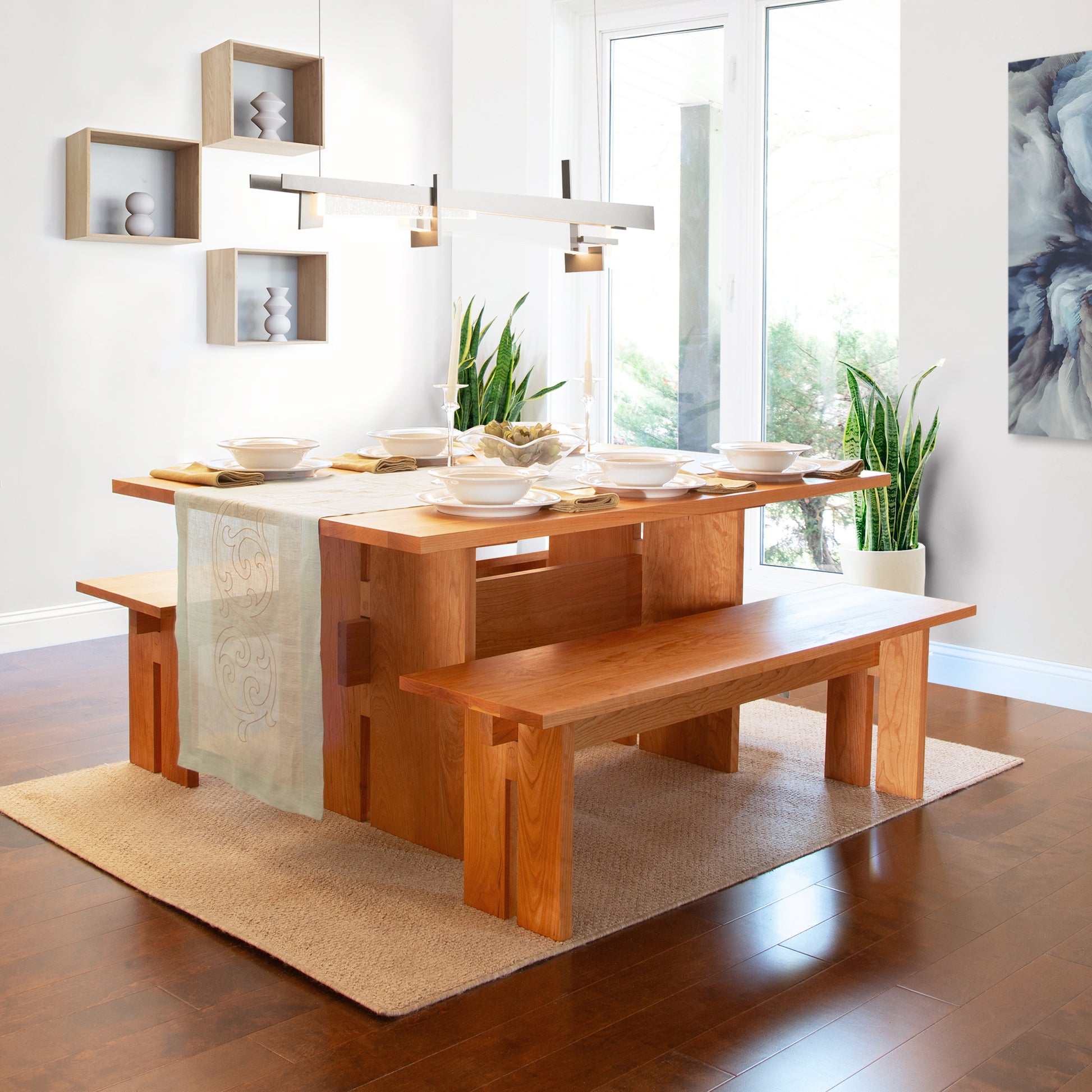 A Vermont Furniture Designs Modern American Dining Table set with matching benches, laid with plates and cups, in a room with hardwood floors, a large window, and modern decor.