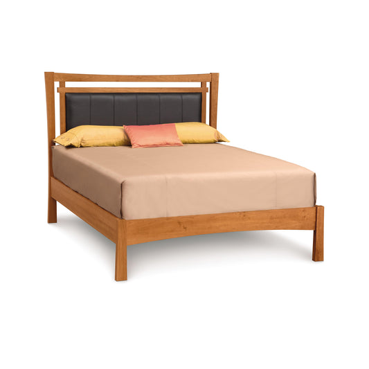 A solid cherry wood frame double Monterey Platform Bed with Upholstered Headboard from Copeland Furniture, dressed with a beige sheet and adorned with two yellow square pillows and one rectangular red pillow, isolated on a white background.