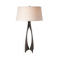 A modern Moreau Table Lamp by Hubbardton Forge with a unique curved wrought iron base and a simple, large cylindrical cream-colored shade, isolated on a white background.