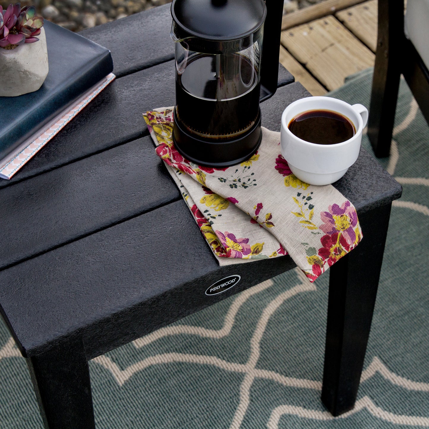 A coffee maker and a cup filled with coffee sit on a weather-resistant POLYWOOD Newport 18" End Table, partially covered by a colorful floral napkin, suggesting a peaceful outdoor coffee break.