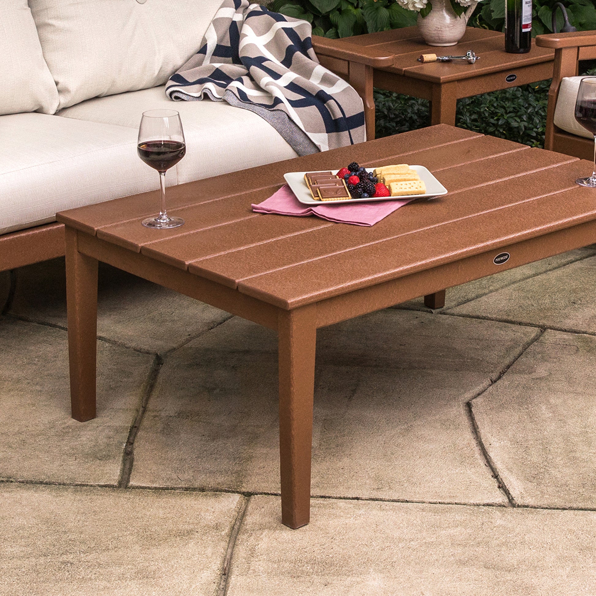 A cozy outdoor setting featuring the POLYWOOD Newport 28" x 42" Coffee Table made from POLYWOOD recycled plastic lumber, holding a glass of red wine, a plate of cookies topped with berries, and a book, between two.