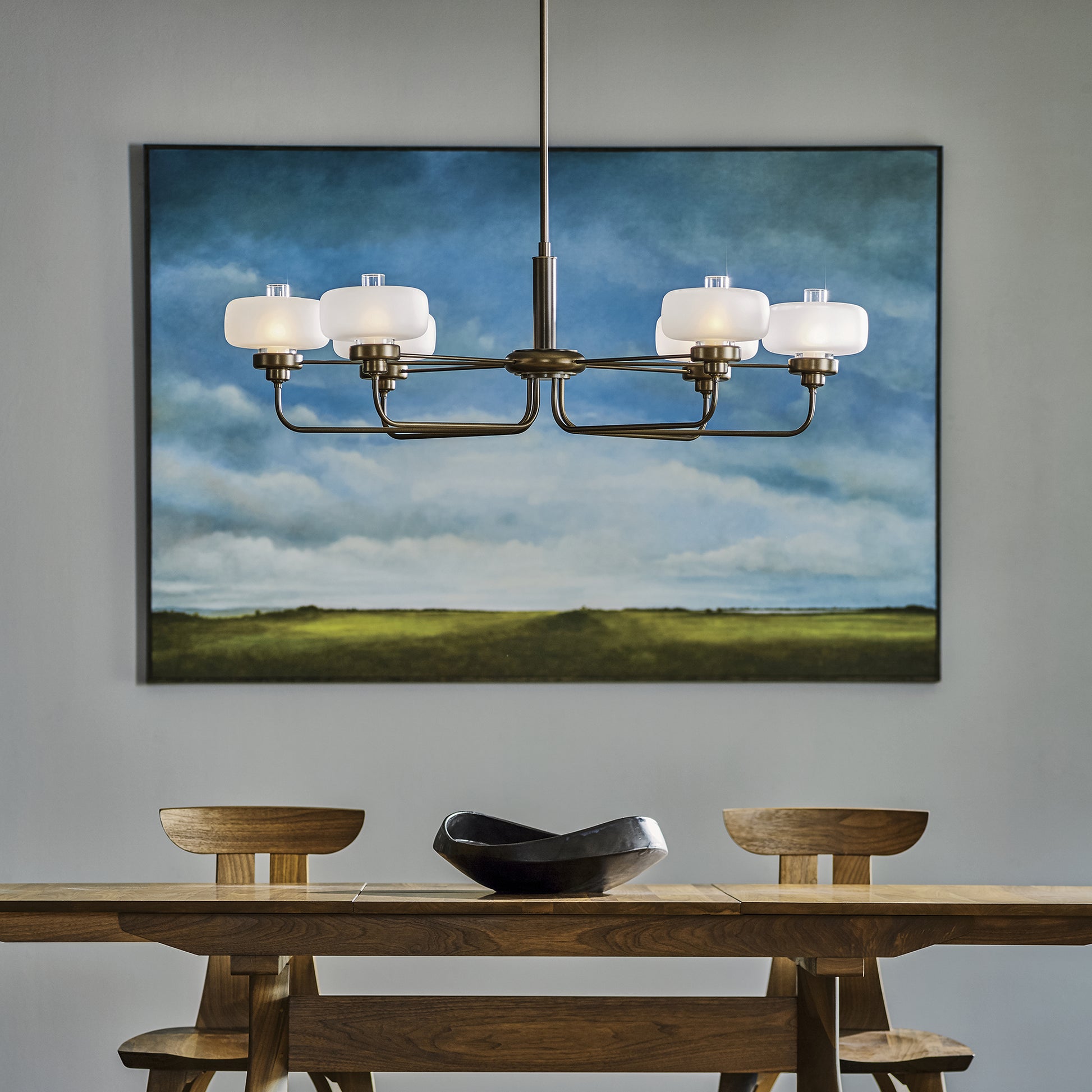 A dining room with a Hubbardton Forge Nola Pendant lighting fixture hanging above a table and chairs, adorned with a beautiful painting.