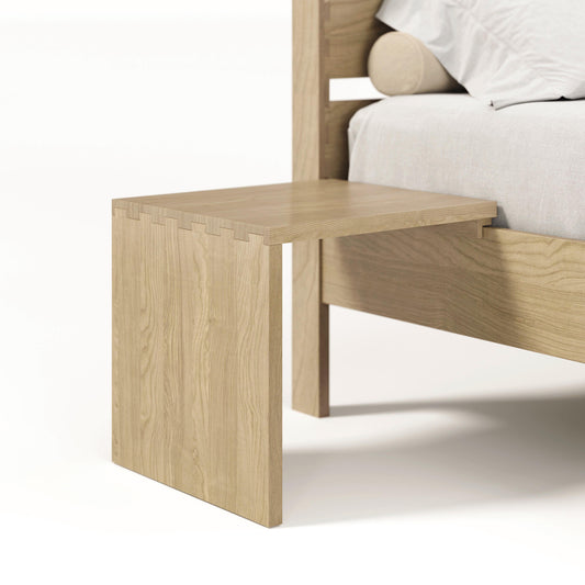 A solid oak hardwood bedside table with a minimalistic design, attached to a bed with a grey upholstered headboard, known as the Oslo Attached Nightstand by Copeland Furniture.