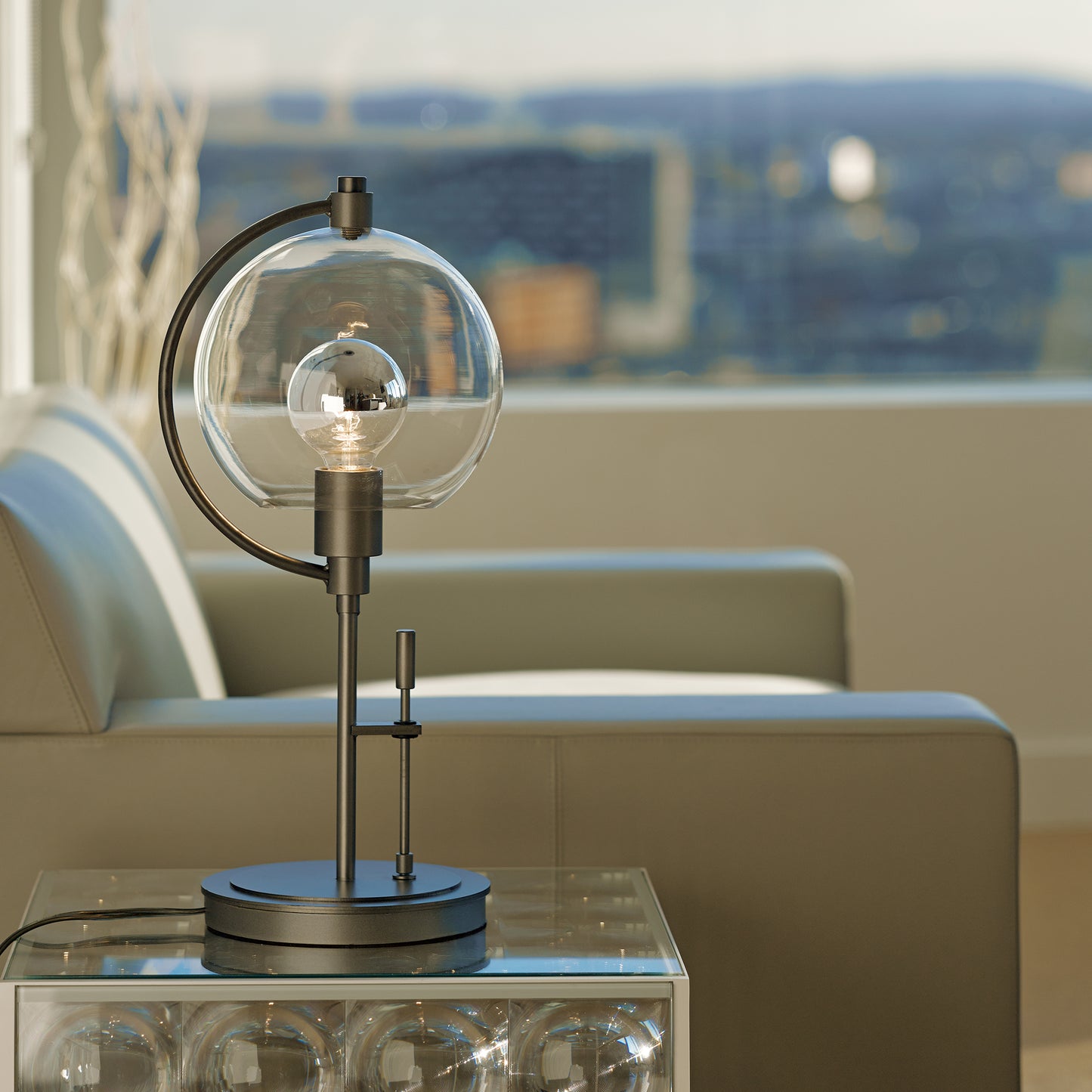 Hubbardton Forge Pluto Table Lamp with a modern metal base and glass globe, displayed on a clear side table.