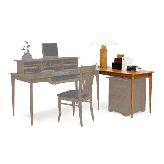 A Copeland Furniture Sarah Return with Keyboard Tray desk setup, crafted from sustainably harvested woods, with an open laptop, a lamp, and office supplies, accompanied by a chair and a side cabinet with books and a plant on a white table.