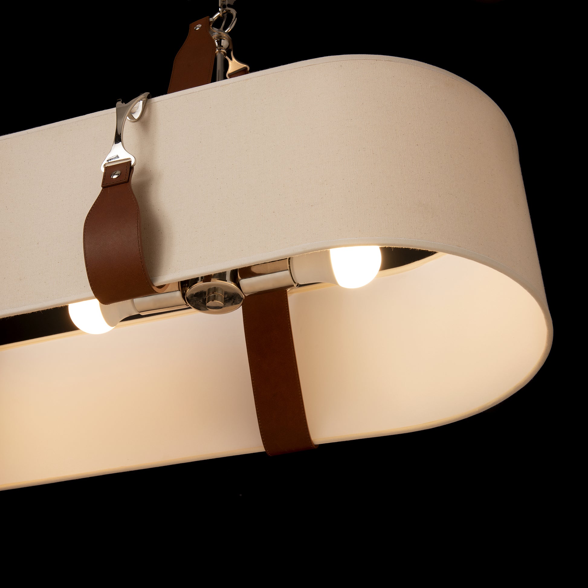 Close-up view of a Hubbardton Forge Saratoga Oval Pendant chandelier with an off-white circular shade held by leather straps, illuminated by several bulbs inside, set against a dark background.