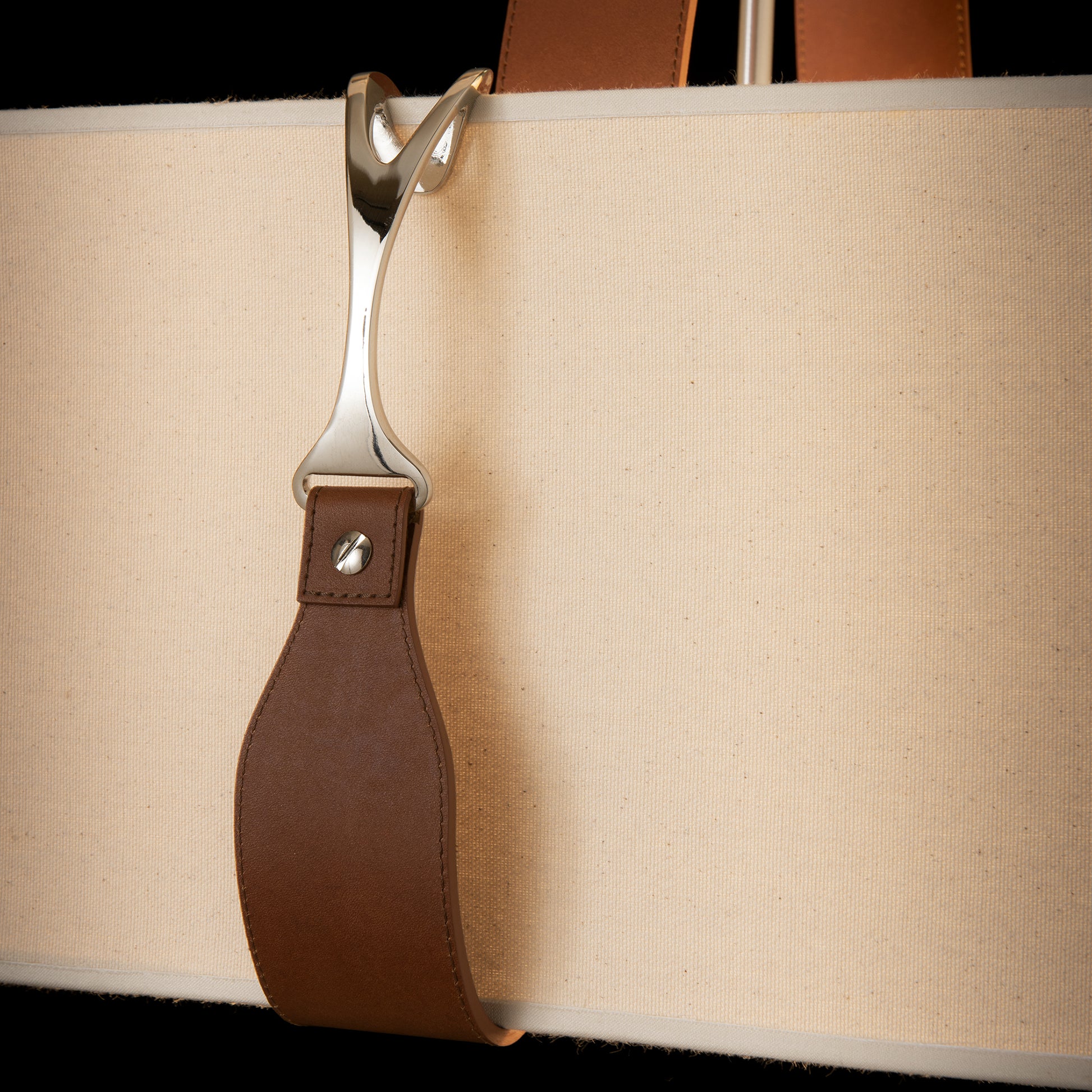 Close-up of a stylish canvas and leather bag, featuring a Saratoga Oval Pendant on the metallic buckle and brown leather straps against a dark background.