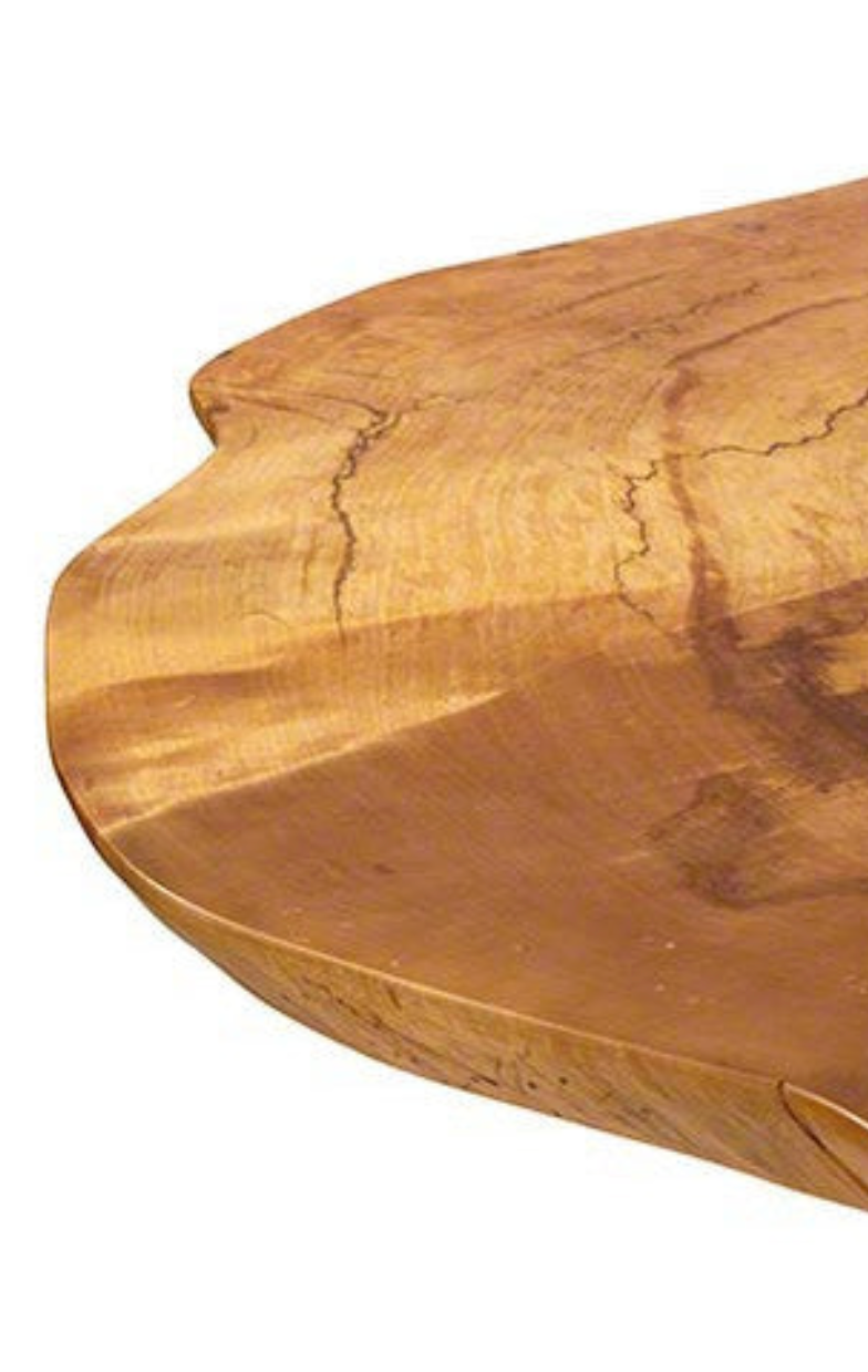 A close-up photo showing the edge of a wooden table with a natural finish. The table features visible wood grain patterns, including lighter and darker tones, with some natural cracks and variations in the surface texture.