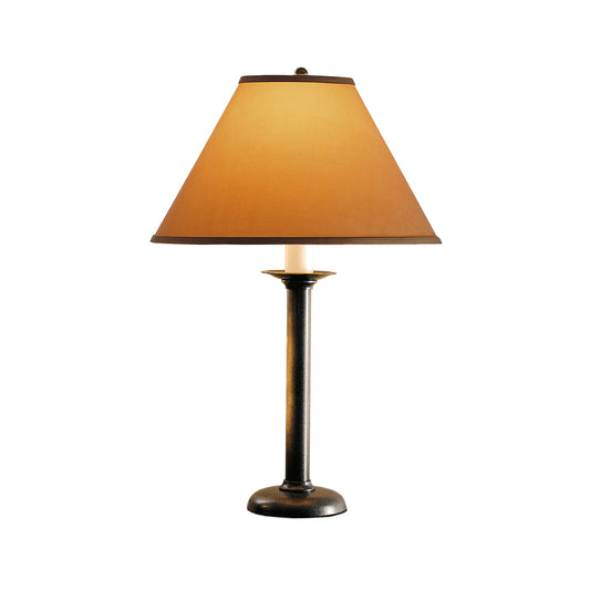 A traditional table lamp with a dark bronze base and a cone-shaped, light orange shade, illuminated. The "Simple Lines Table Lamp" by Hubbardton Forge is centered on a plain white background.