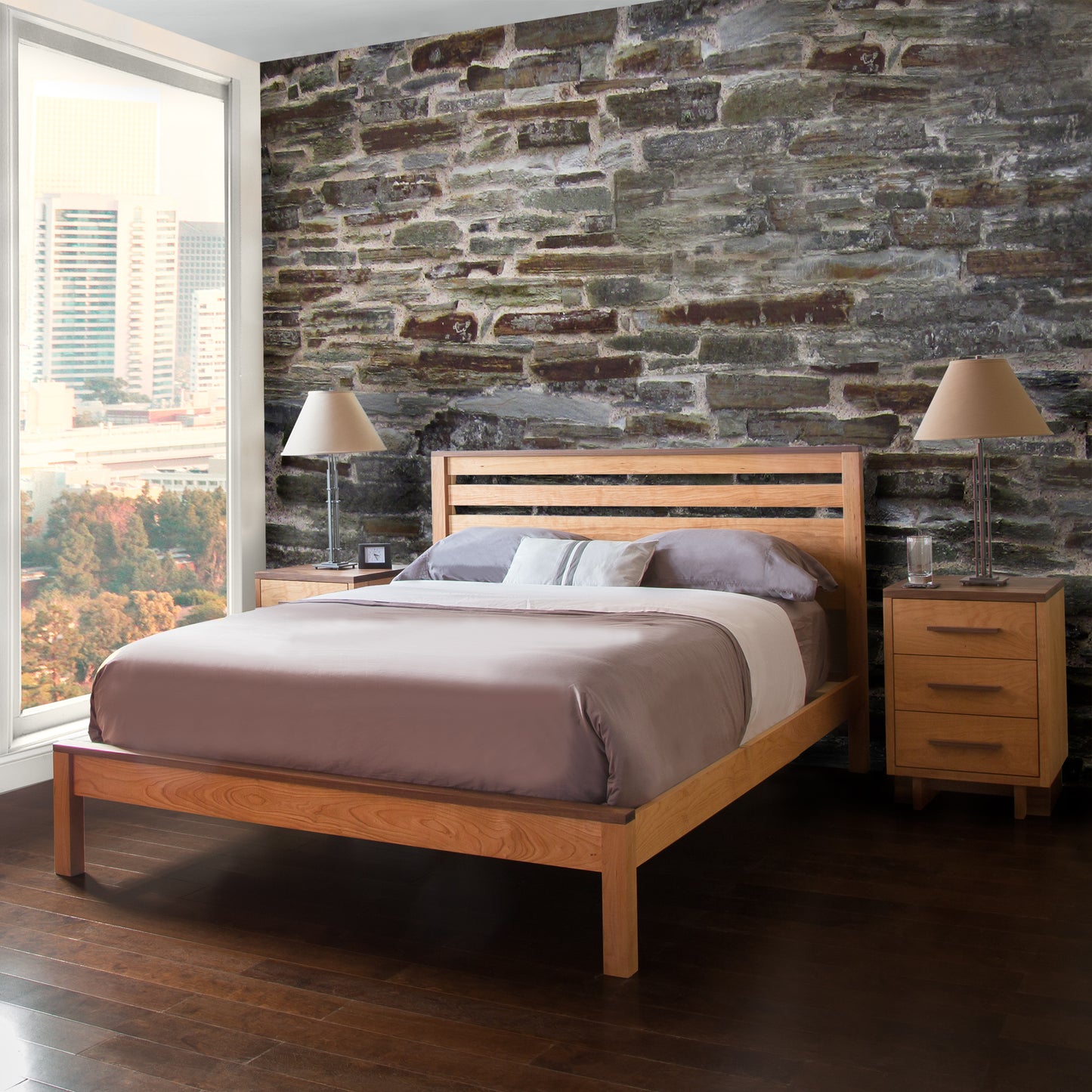 A modern bedroom with a solid Vermont Furniture Designs Skyline Panel Bed, matching nightstands, and beige bedding against a stone accent wall, with a cityscape visible through the window.