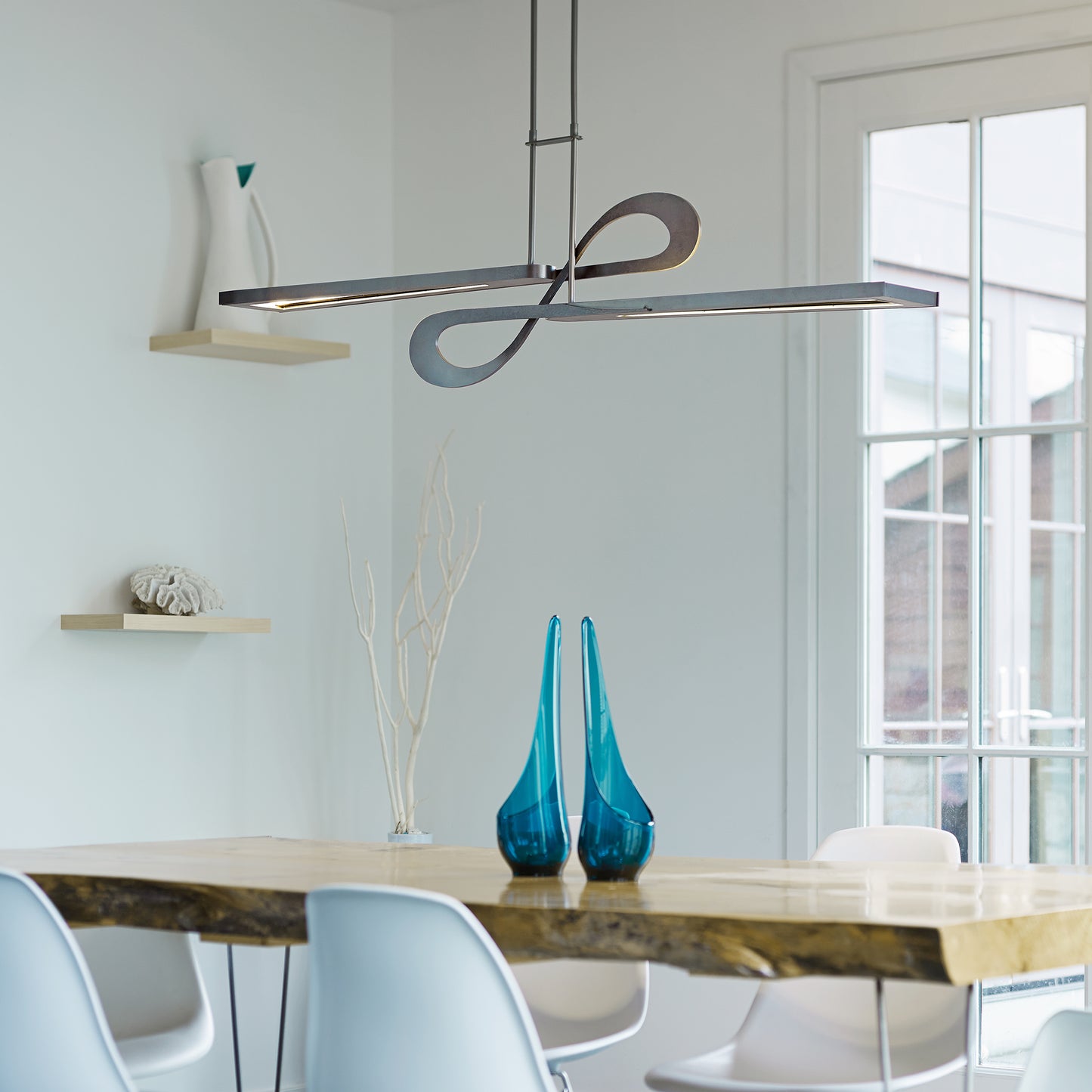 Elegant wooden dining table with white chairs, highlighted by a Hubbardton Forge Switchback Pendant above; minimal decor in view.