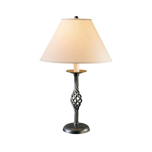 Hubbardton Forge Twist Basket Table Lamp with a hand-forged iron base and a tapered beige lampshade, isolated on a white background.