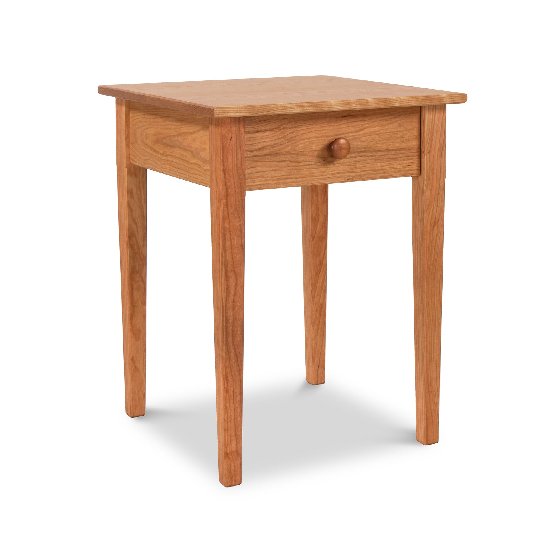 A Vermont Shaker Bedside Table from Maple Corner Woodworks, with a single drawer and a round knob, standing on four slender legs, isolated against a white background.