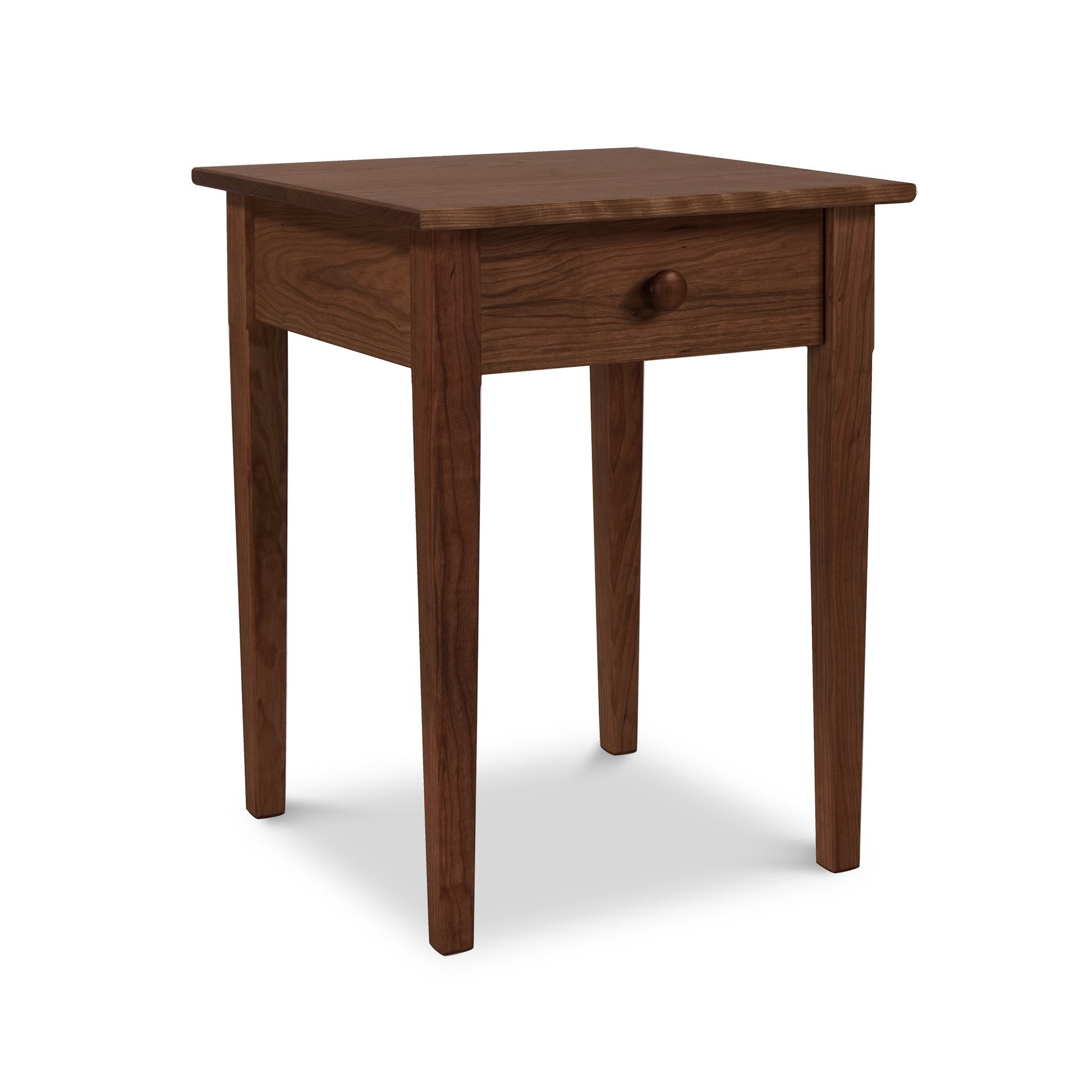 A Vermont Shaker Bedside Table, crafted from sustainably harvested solid woods by Maple Corner Woodworks, featuring a single drawer, stands against a plain white background. The table boasts a smooth top and slightly tapered legs.